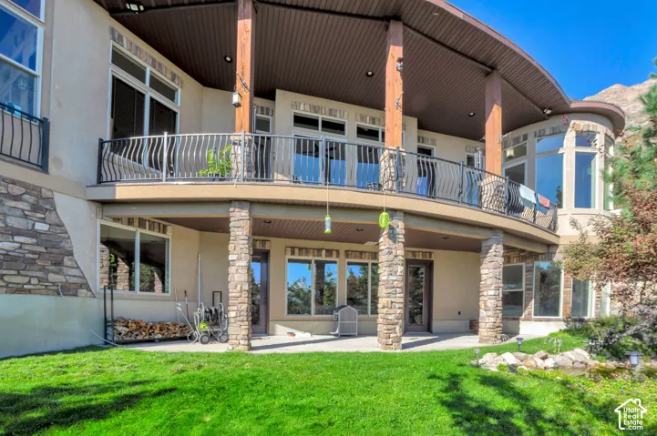 Curved upper balcony with black metal railing, stone-clad pillars, a lower patio, and a manicured lawn