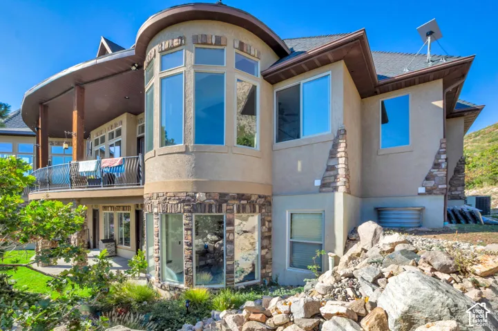 Stucco exterior with stone accents, round turret with expansive windows, elevated balcony with metal railing, covered patio, and multi-level landscaping