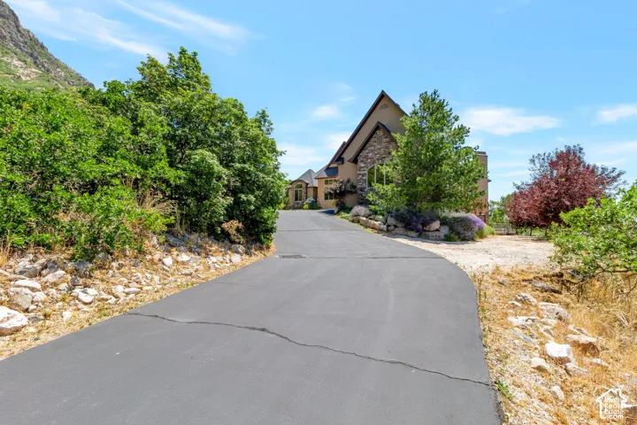 Curved asphalt driveway ascending to a stucco and stone-clad residence, featuring a peaked roofline and mature landscaping