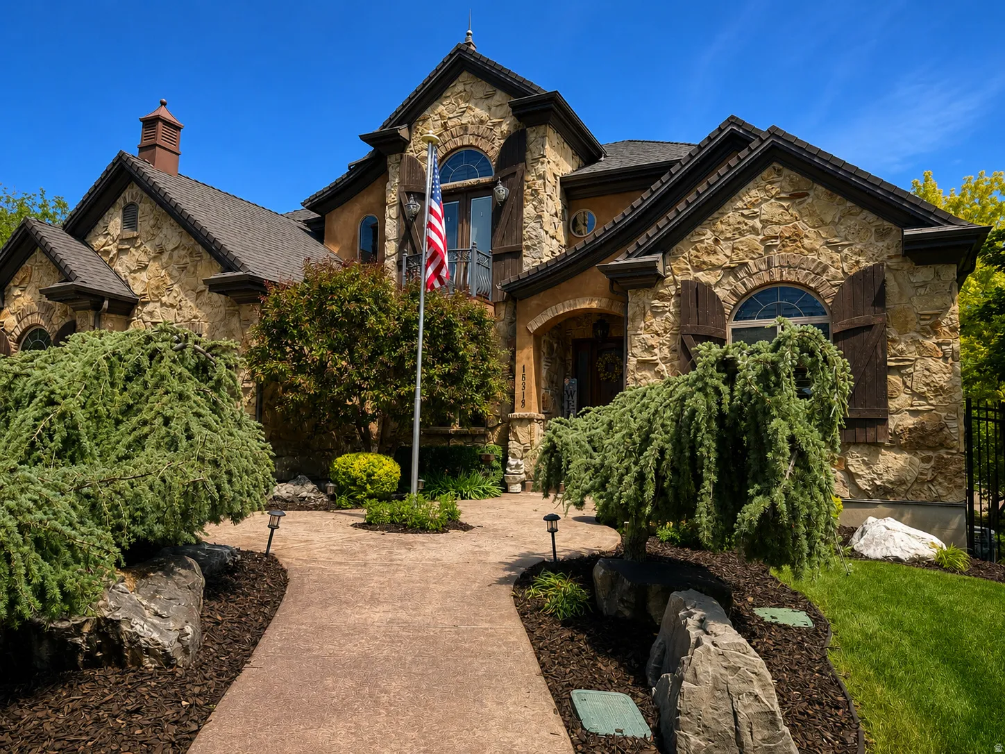 Stone facade residence featuring a prominent arched entry, decorative window shutters, and a mature landscaped front yard with a winding pathway