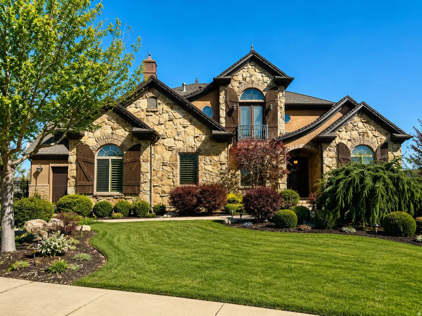 Stone and stucco exterior featuring a prominent arched entry, dark wood-tone shutters, and an upper-level balcony with iron railing