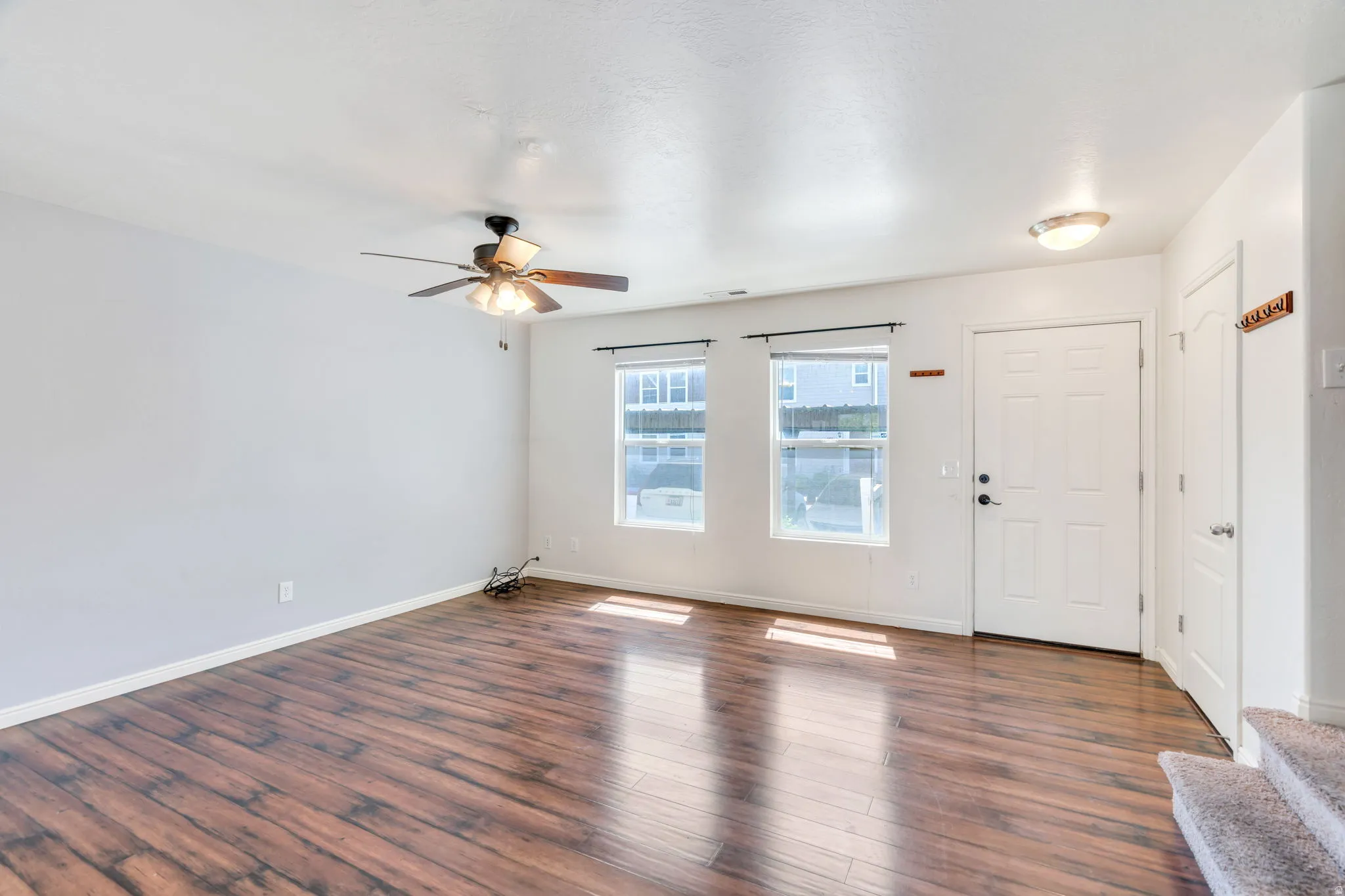 Spacious room featuring wood-finish flooring, two windows with blinds, a ceiling fan, a white panel door, and white baseboards