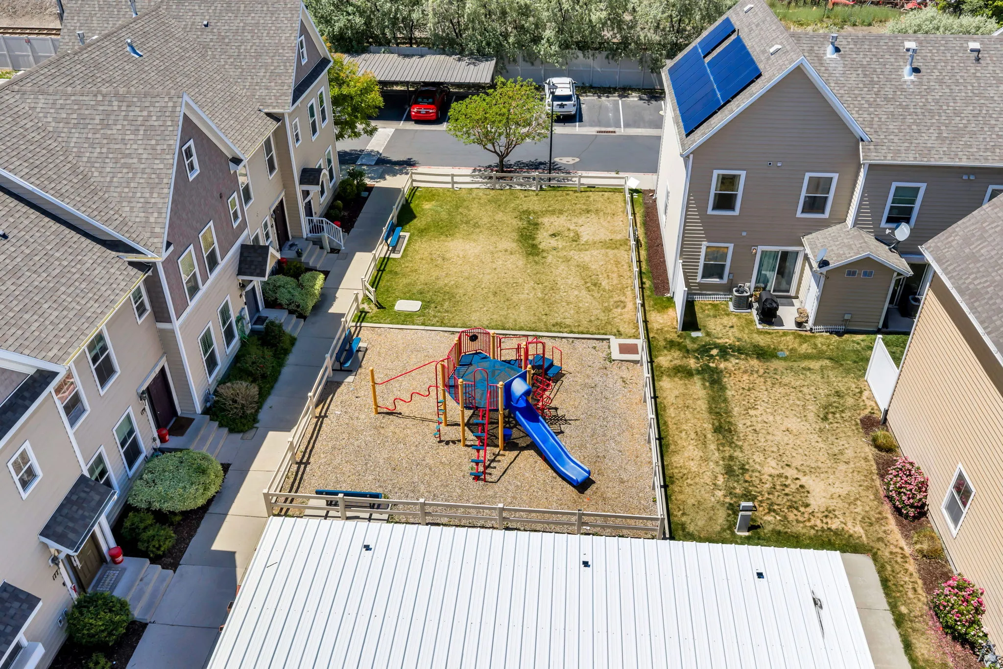 Community courtyard featuring a fenced playground with a slide and climbing structure, surrounded by a grassy area and mature trees