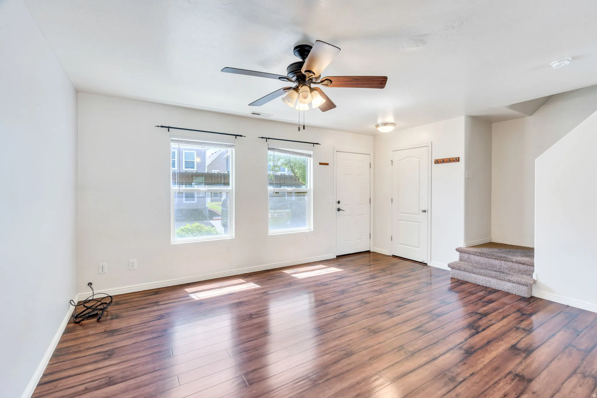 Spacious room featuring wood-finish flooring, two windows with blinds, a ceiling fan with integrated lighting, and two white doors