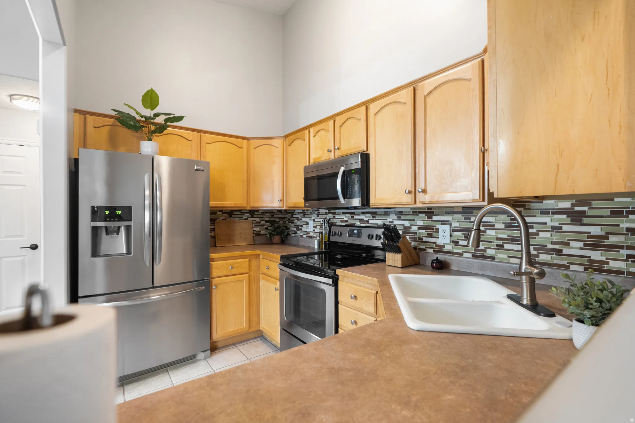 Kitchen with stainless steel appliances, light wood finish cabinetry, light tile patterned flooring, a high ceiling, and backsplash