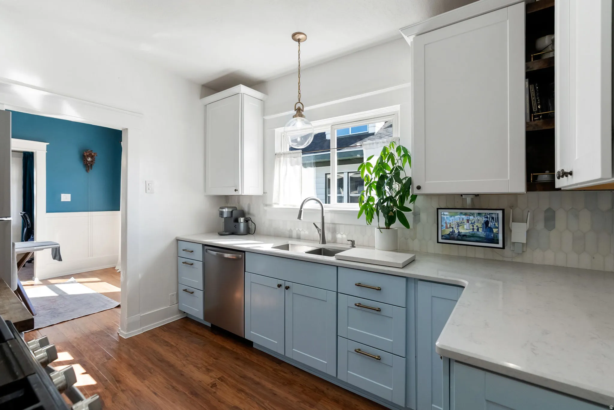 Kitchen with light stone counters, dark wood finished floors, stainless steel dishwasher, hanging light fixtures, and a wainscoted wall