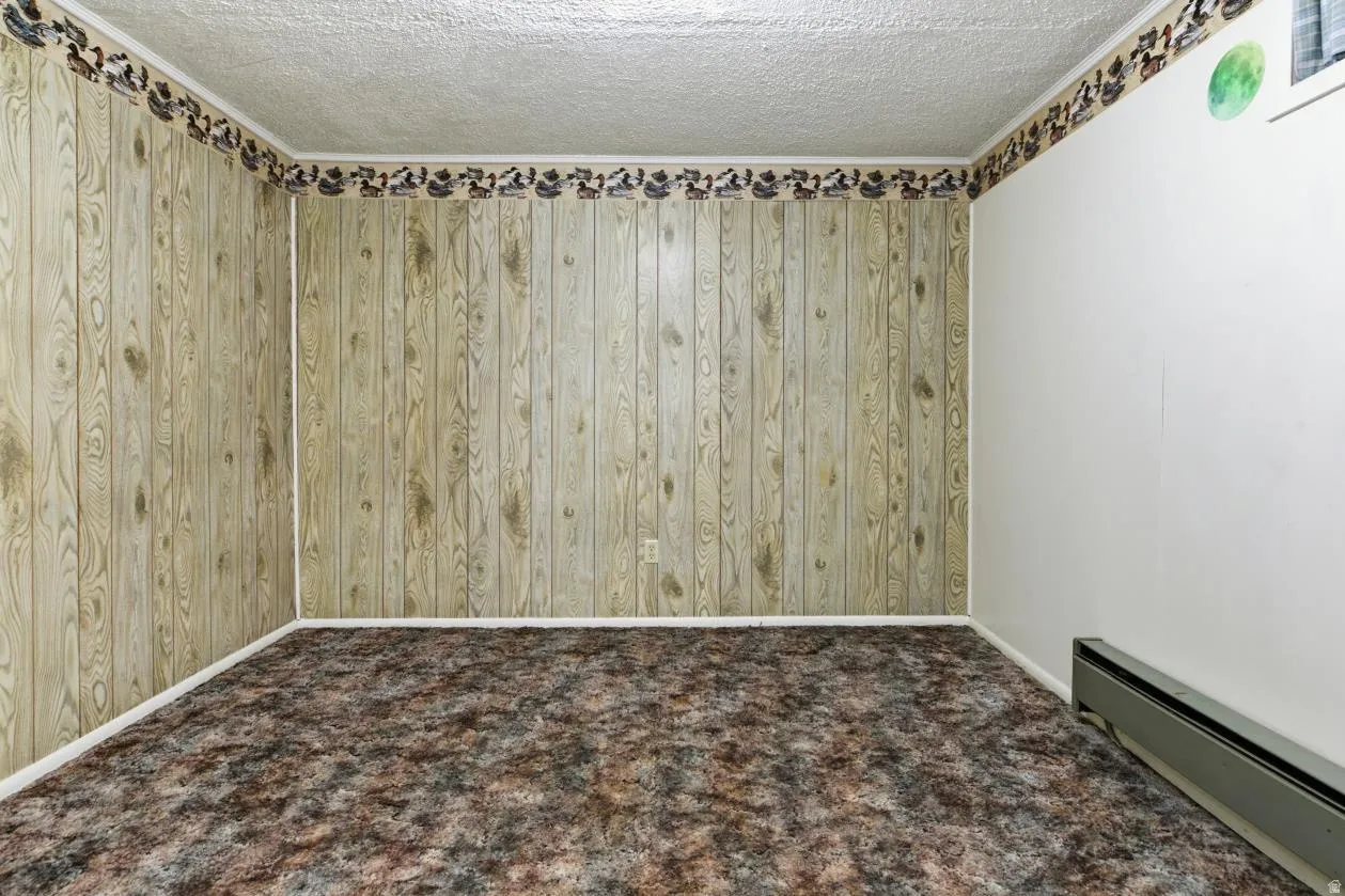 Carpeted empty room featuring a baseboard heating unit, a textured ceiling, and wood walls