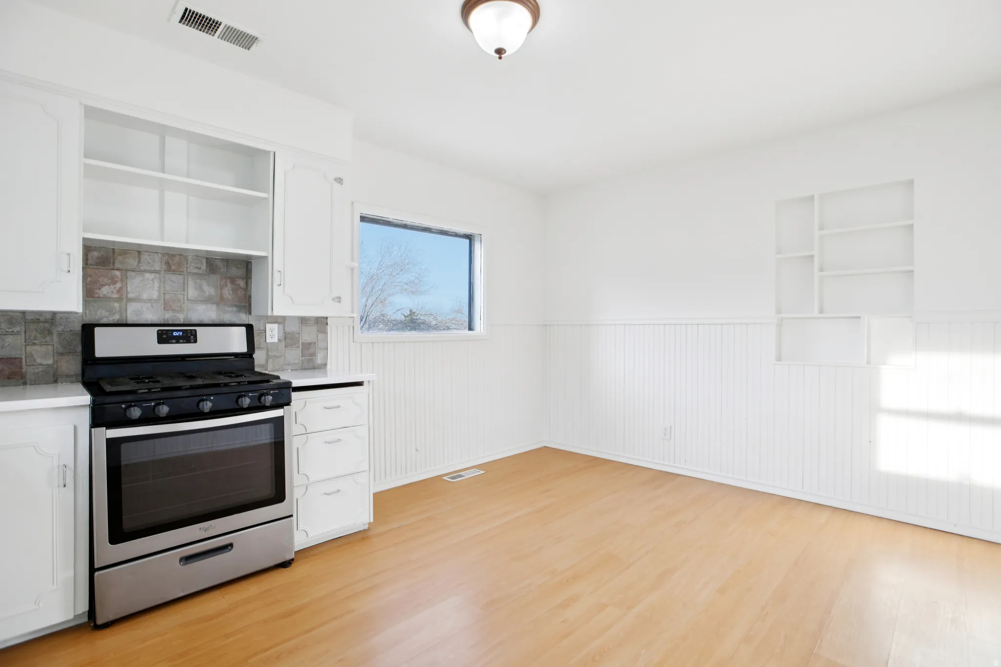 Kitchen with open shelves, stainless steel gas stove, white cabinetry, wainscoting, and light countertops