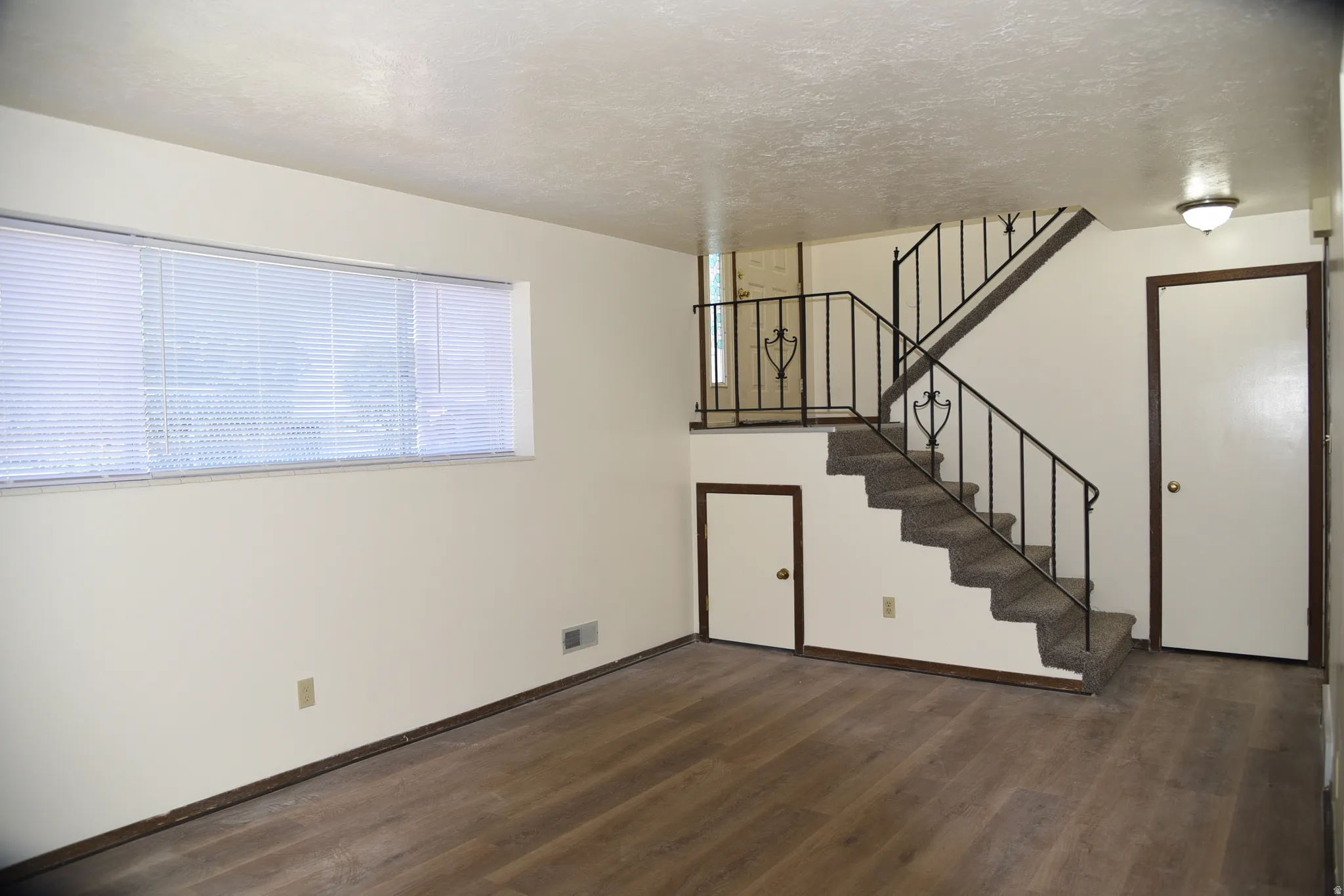 Living space featuring wood-finish flooring, a multi-level staircase with wrought iron railing, a wall-mounted light fixture, and a large window with blinds