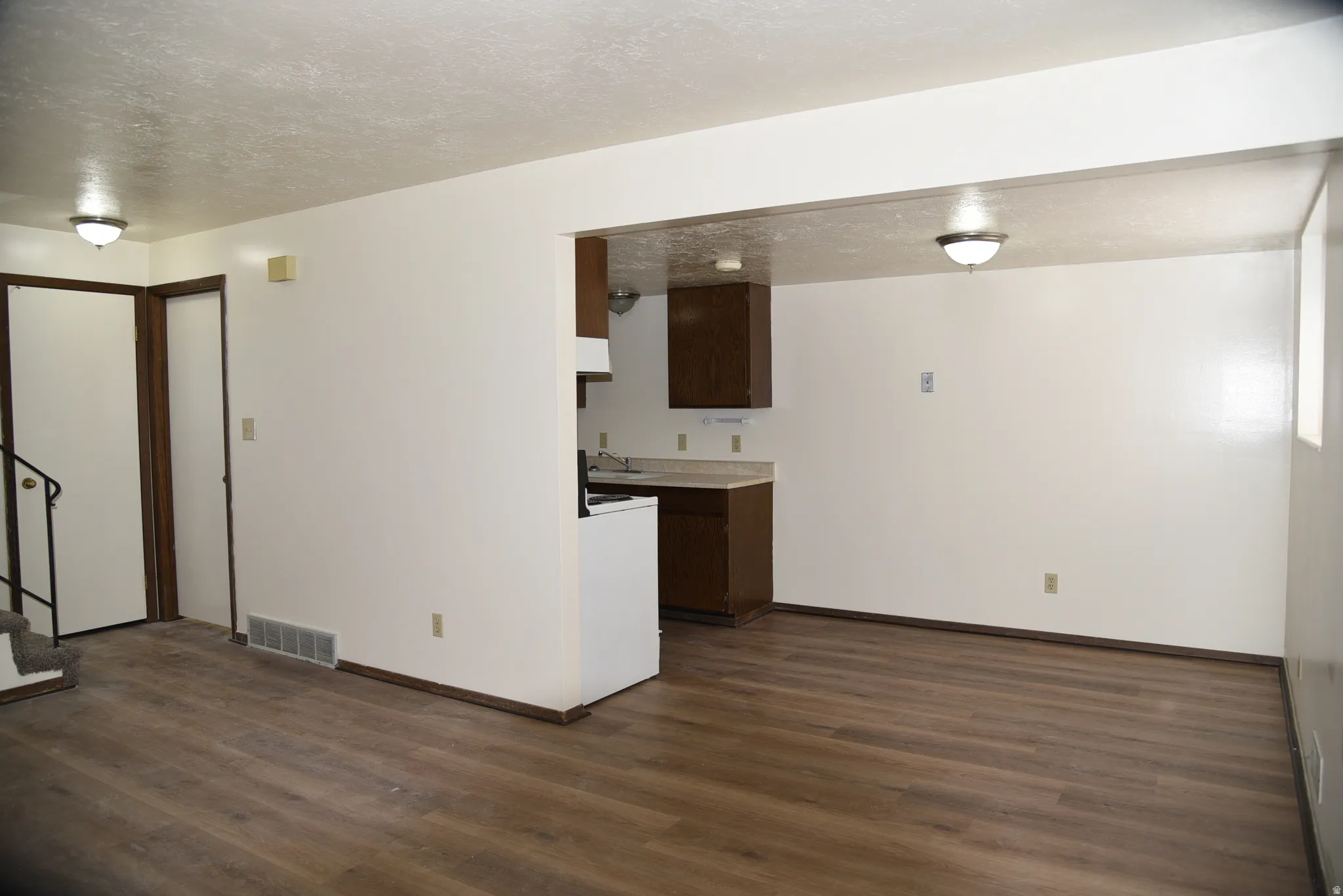Open-concept living area featuring wood-finish flooring, white walls, and a visible kitchen area with brown cabinetry and light-colored countertops