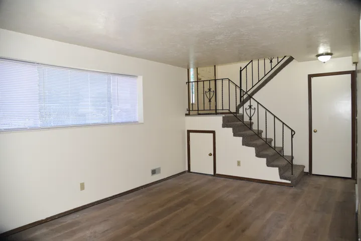 Living space featuring wood-finish flooring, a multi-level staircase with wrought iron railing, a wall-mounted light fixture, and a large window with blinds