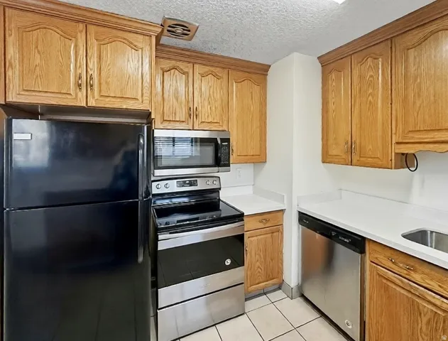 Kitchen with stainless steel appliances, a textured ceiling, light tile patterned floors, light stone countertops, and wood finish cabinetry