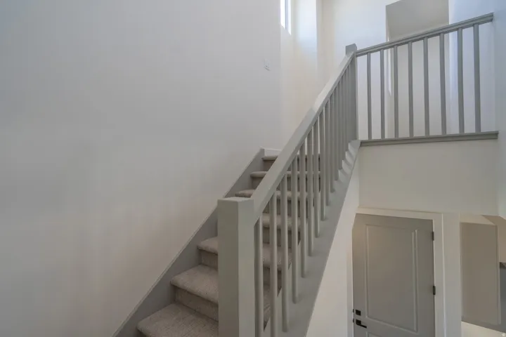 Carpeted staircase featuring a light gray handrail and balusters, white walls, and a light-colored door with black hardware