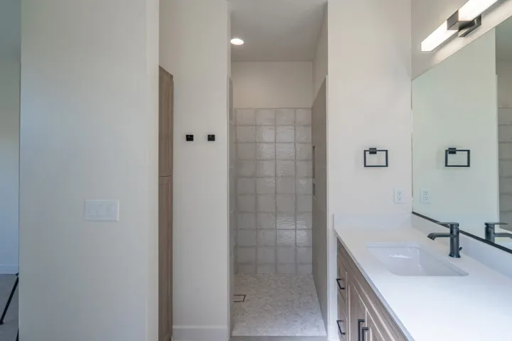 Modern bathroom featuring a walk-in shower with square wall tiling and pebble flooring, a light wood-finish vanity with a white countertop and integrated sink, a large mirror, and contemporary lighting