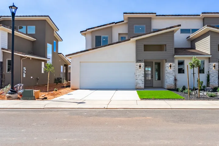 Contemporary facade featuring a white garage door, stacked stone accents, decorative exterior lighting, and a low-maintenance artificial turf lawn