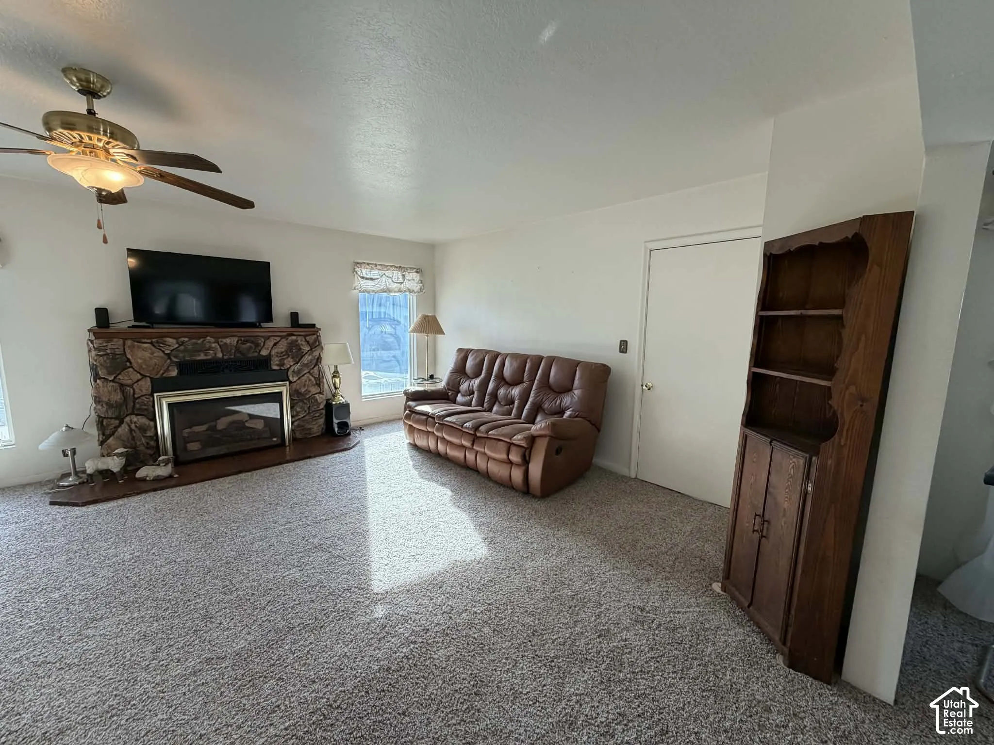 Living area featuring light carpet, a stone fireplace, ceiling fan, and a textured ceiling