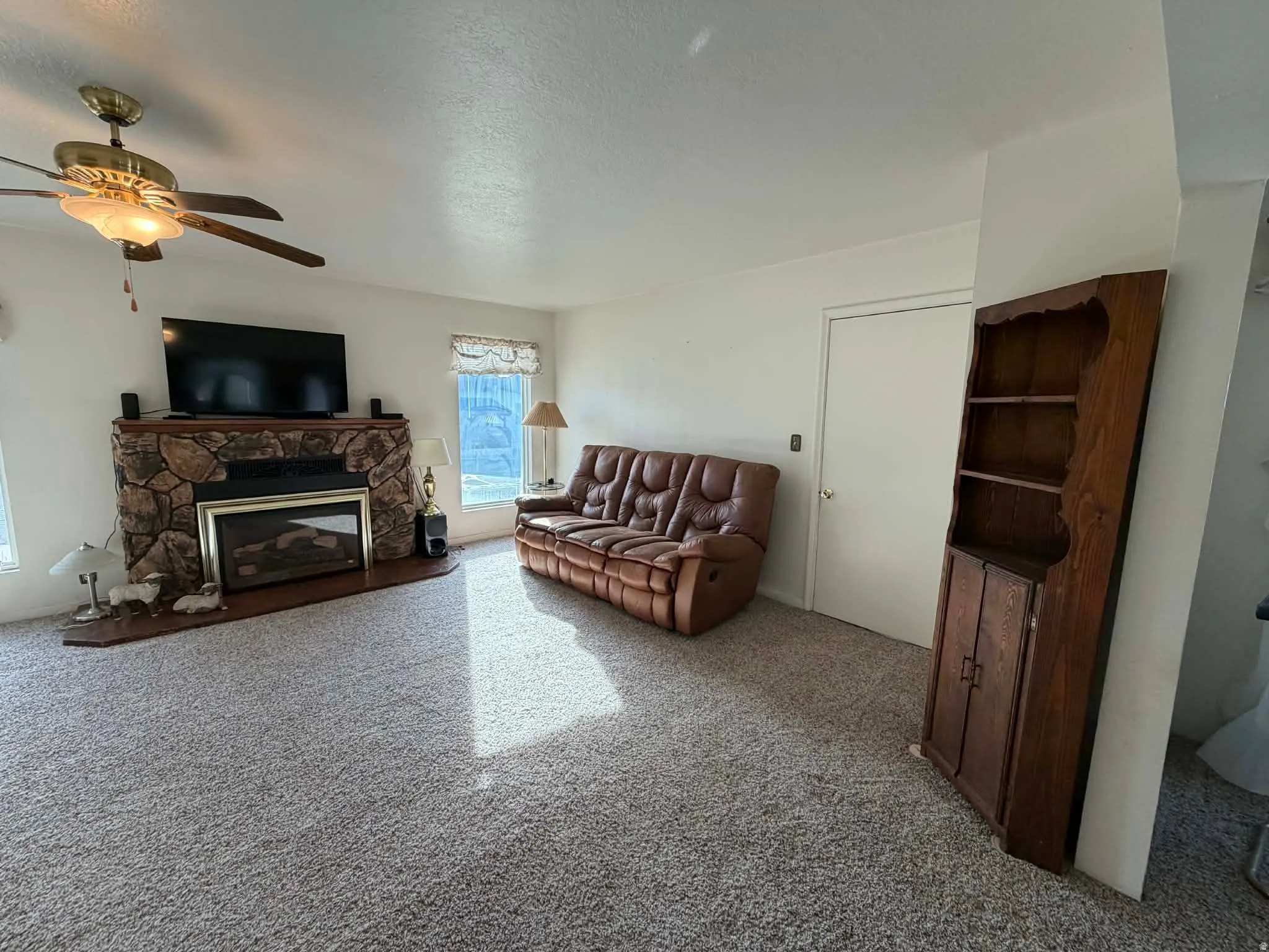 Living area featuring light carpet, a stone fireplace, ceiling fan, and a textured ceiling