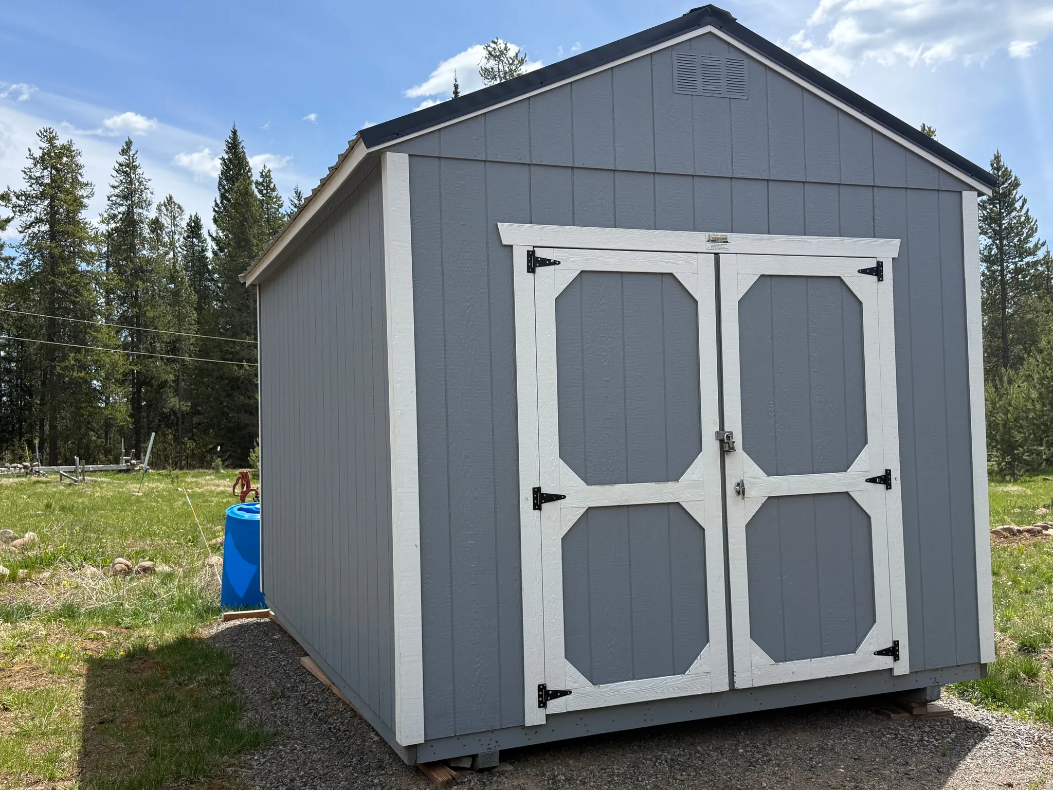Gable-roof storage shed featuring gray siding with white trim, double barn-style doors with black hardware, and an upper gable vent