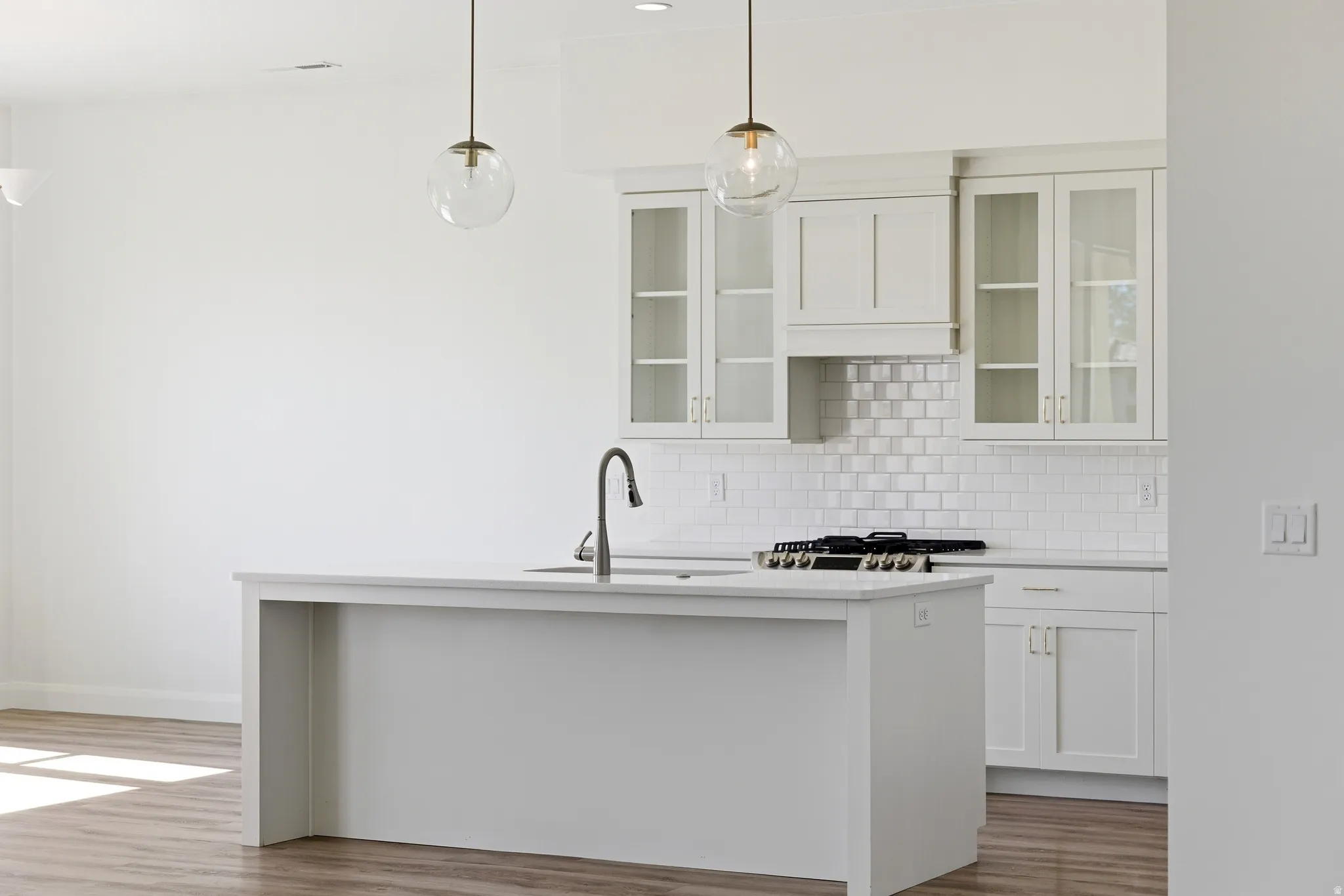 Kitchen featuring white cabinetry, hanging light fixtures, glass fronted cabinets, tasteful backsplash, and a center island with sink