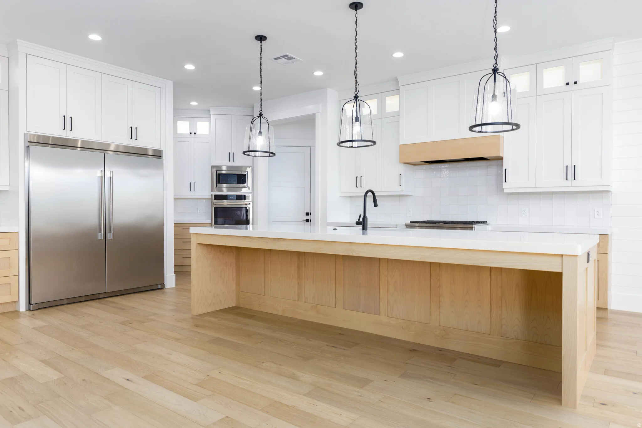 Kitchen with built in appliances, glass insert cabinets, a center island with sink, light wood-style flooring, and two tone cabinetry