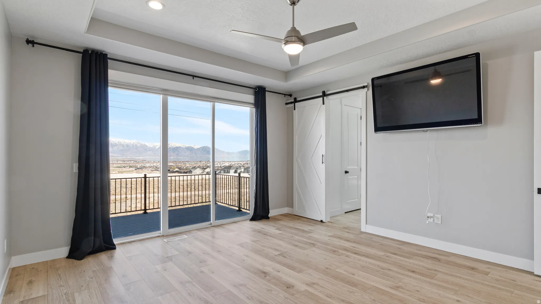 Unfurnished bedroom with a barn door, light wood-style flooring, access to outside, a ceiling fan, and a tray ceiling