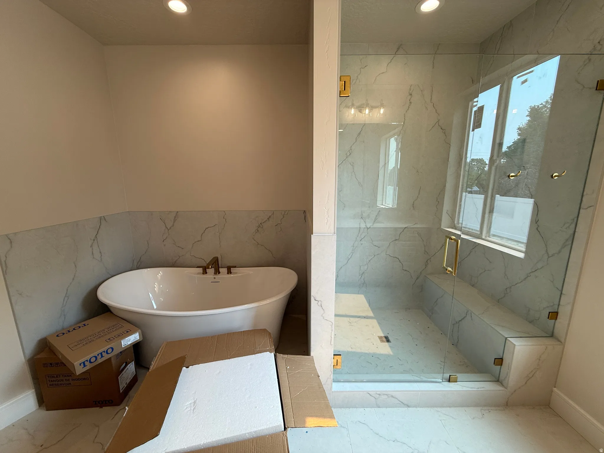 Freestanding soaking tub with bronze-finish fixtures, complemented by a spacious glass-enclosed shower featuring marble-look tile, a built-in bench, and a window