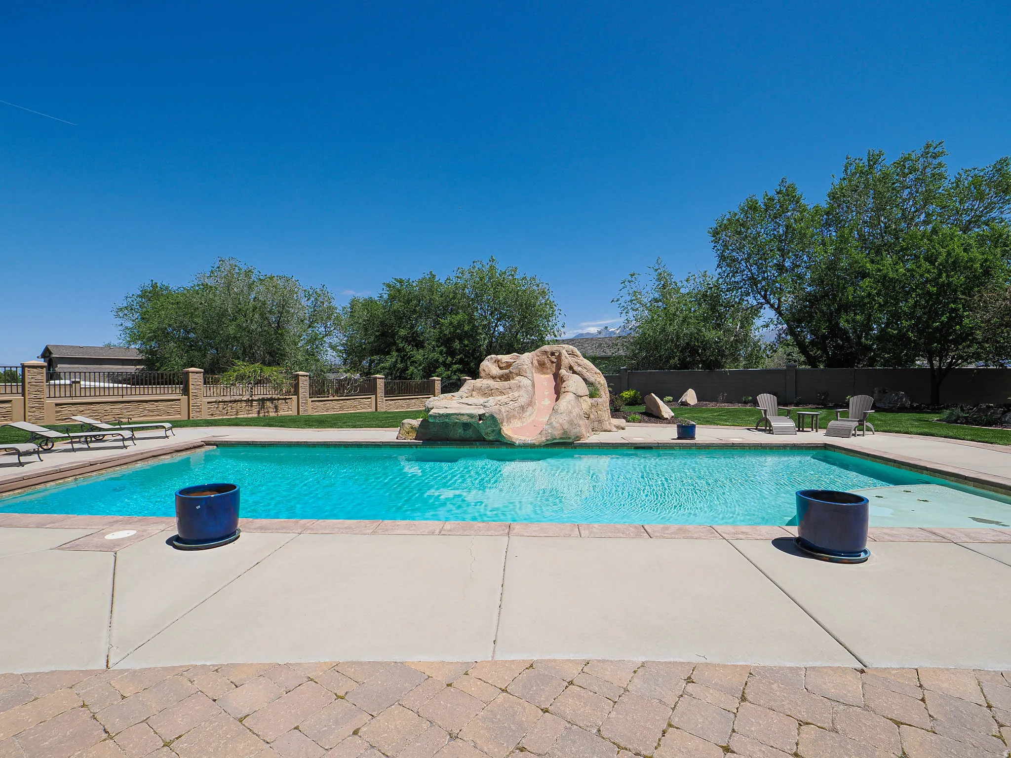 Expansive swimming pool featuring a rock-formation slide, surrounded by a concrete patio and a paver stone deck