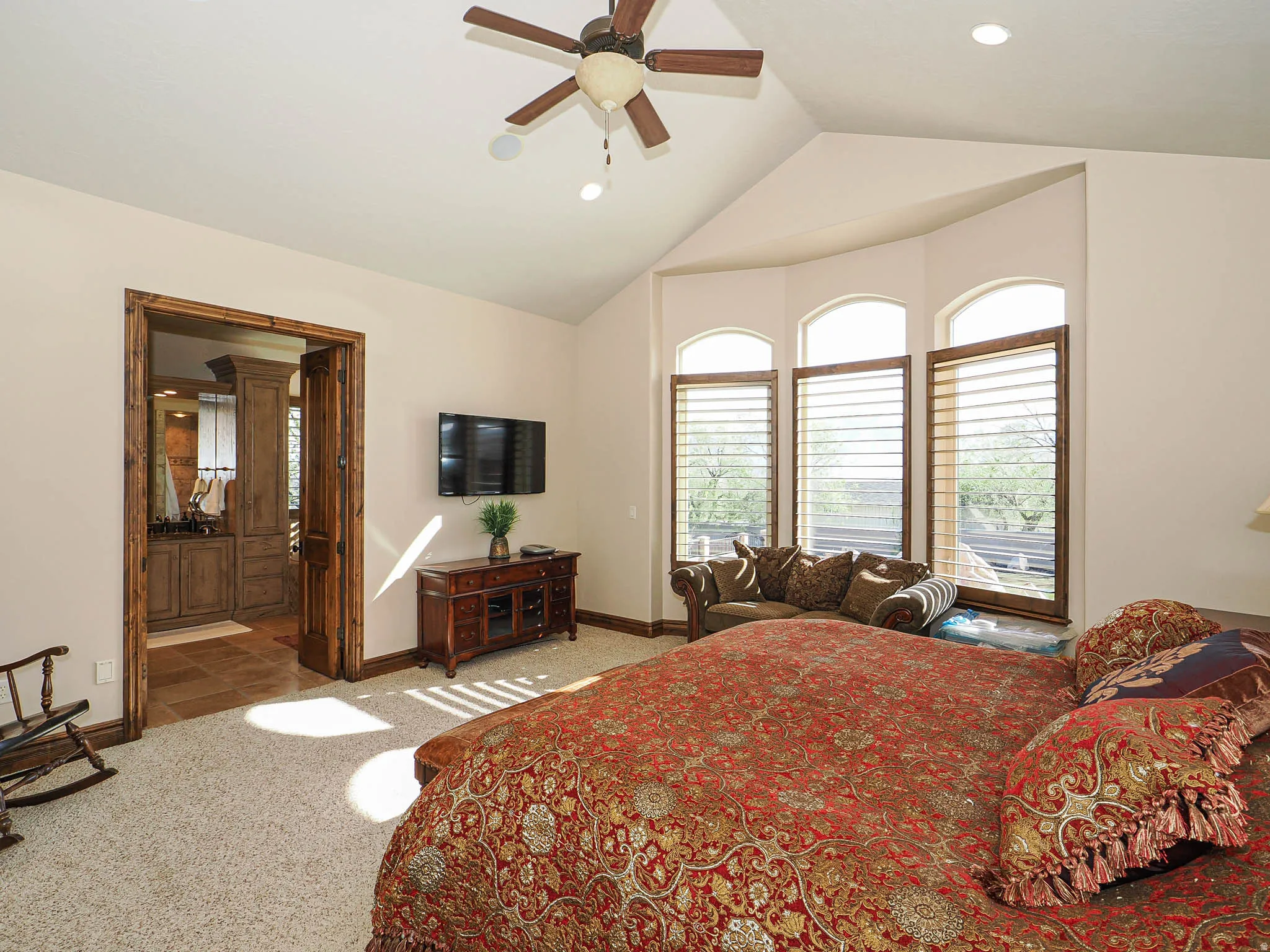 Vaulted ceiling featuring a ceiling fan and recessed lighting, plush carpet flooring, a bay window with arched transoms and plantation shutters, and an adjacent bathroom with wood-finish cabinetry and tiled flooring