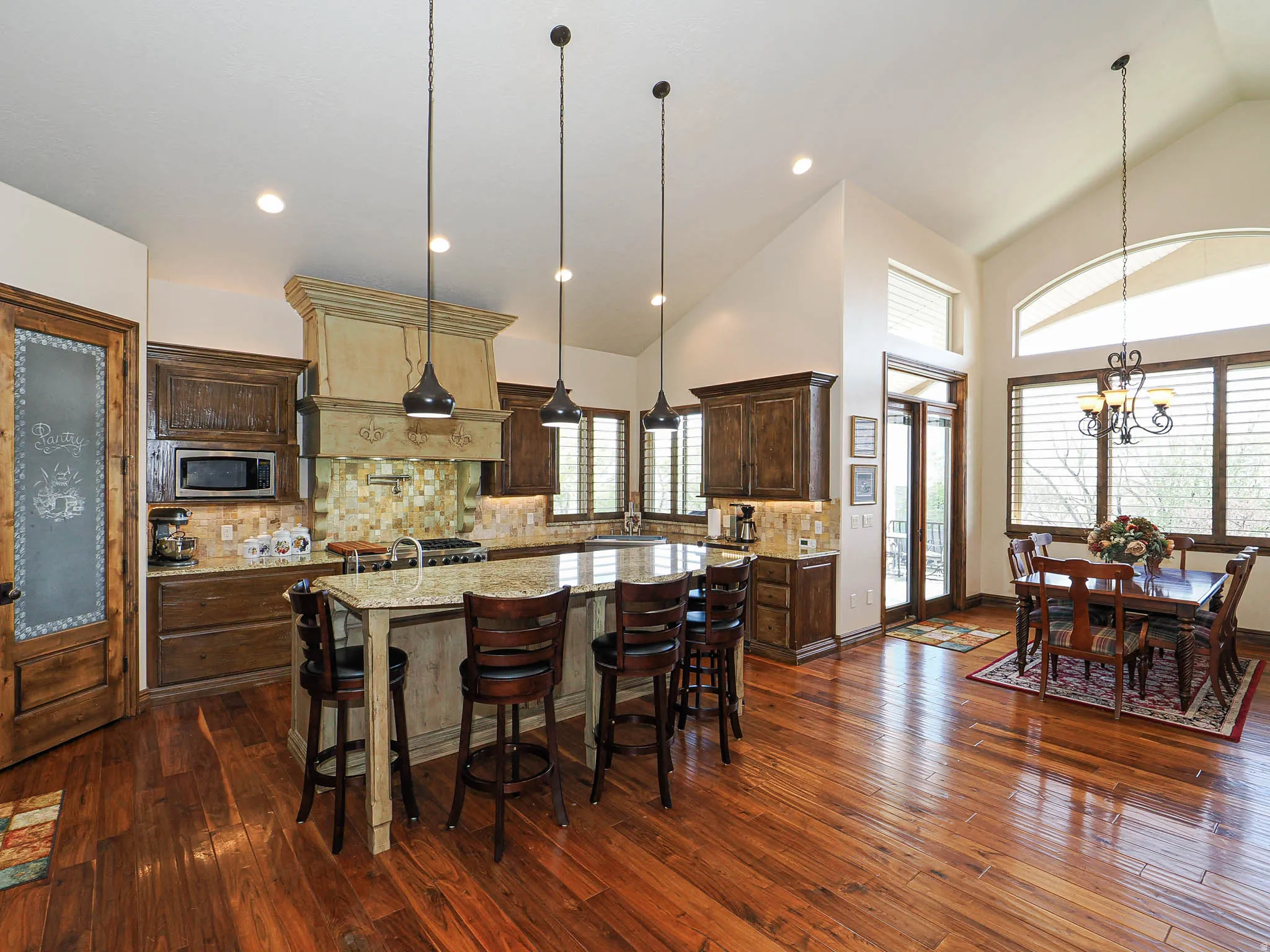 Gourmet kitchen and dining area featuring rich wood-finish flooring, a large granite island with seating, dark wood cabinetry, a decorative range hood, and a pantry with a frosted glass door