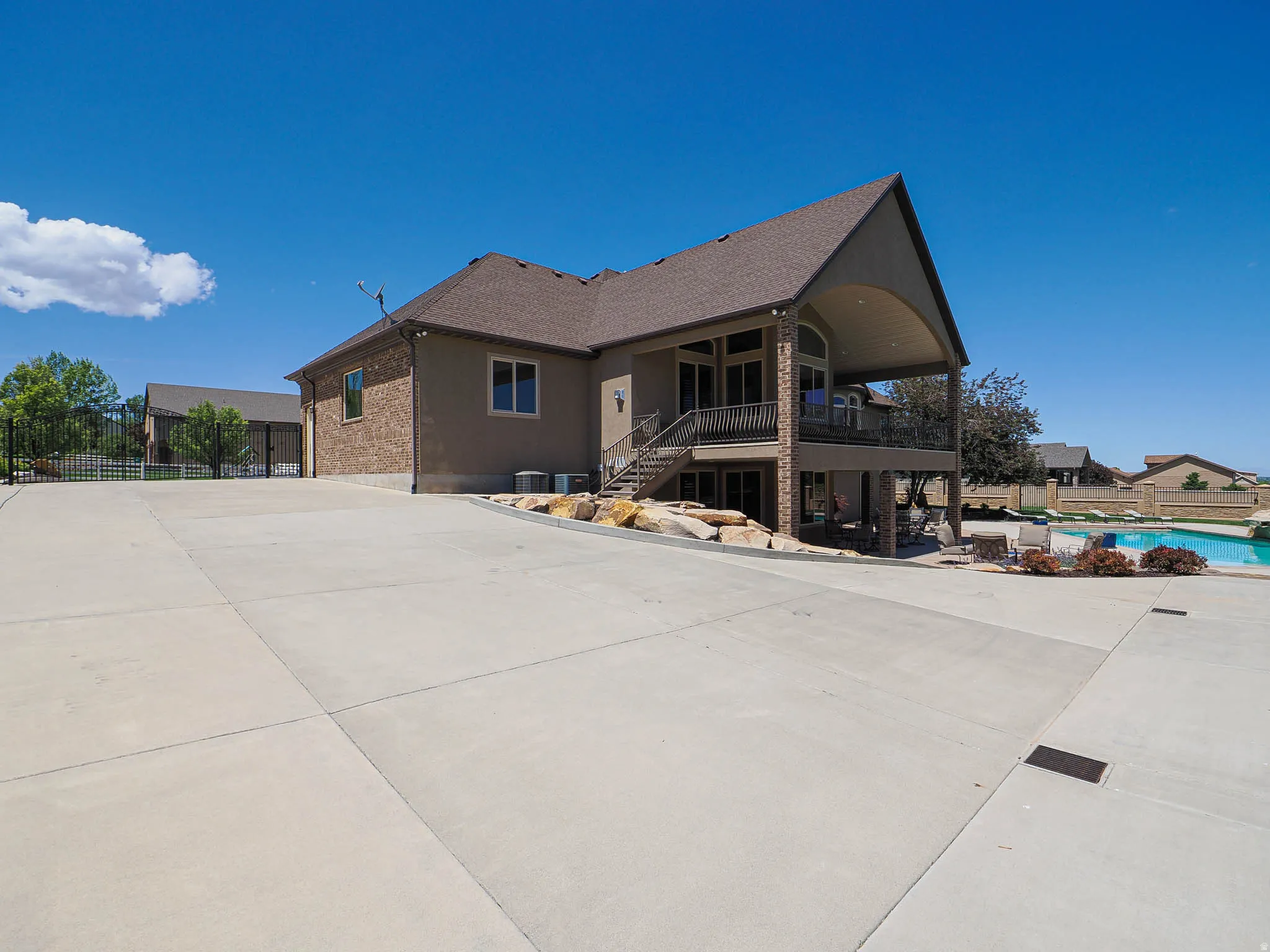 Spacious concrete driveway leading to a multi-level residence featuring a stucco and brick exterior, covered upper and lower patios, and an outdoor swimming pool