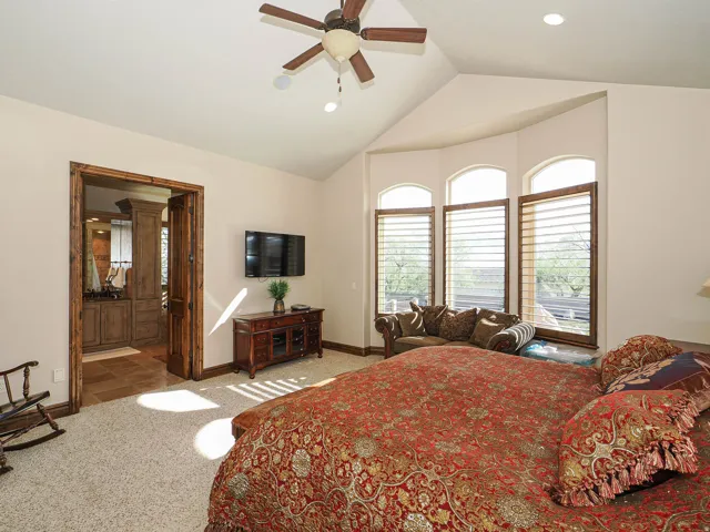 Vaulted ceiling featuring a ceiling fan and recessed lighting, plush carpet flooring, a bay window with arched transoms and plantation shutters, and an adjacent bathroom with wood-finish cabinetry and tiled flooring