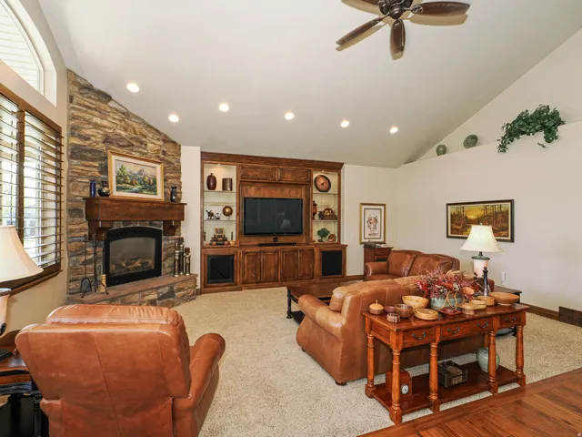 Living area featuring a stone-clad fireplace with a wood mantle, a built-in entertainment center with display shelving, vaulted ceilings, recessed lighting, and wood-finish window shutters