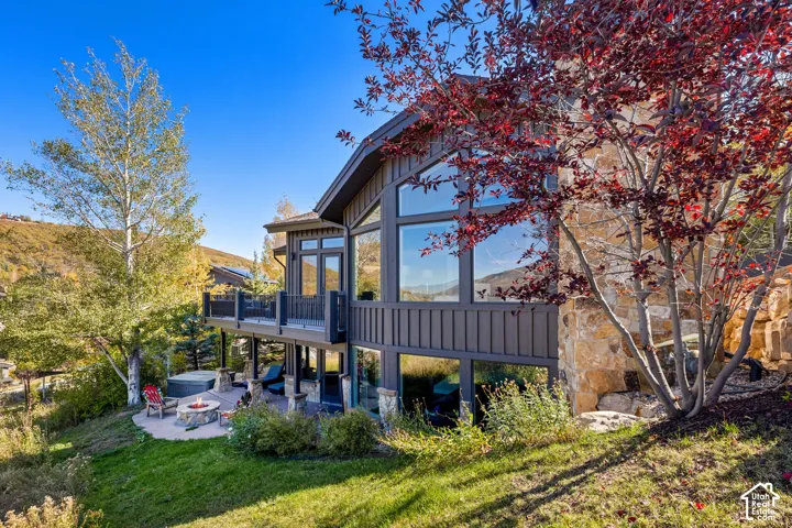Exterior featuring vertical siding, multi-story windows, stone chimney, elevated deck with railing, and a stone-paver patio