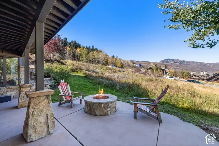 Covered patio featuring a concrete floor, stone-clad support pillars, and a circular stone fire pit, set against a mountainous landscape