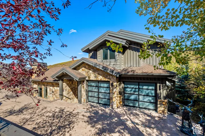 Multi-level exterior featuring vertical board-and-batten siding, stone masonry facade, dual modern garage doors with frosted glass panels, and an expansive concrete driveway