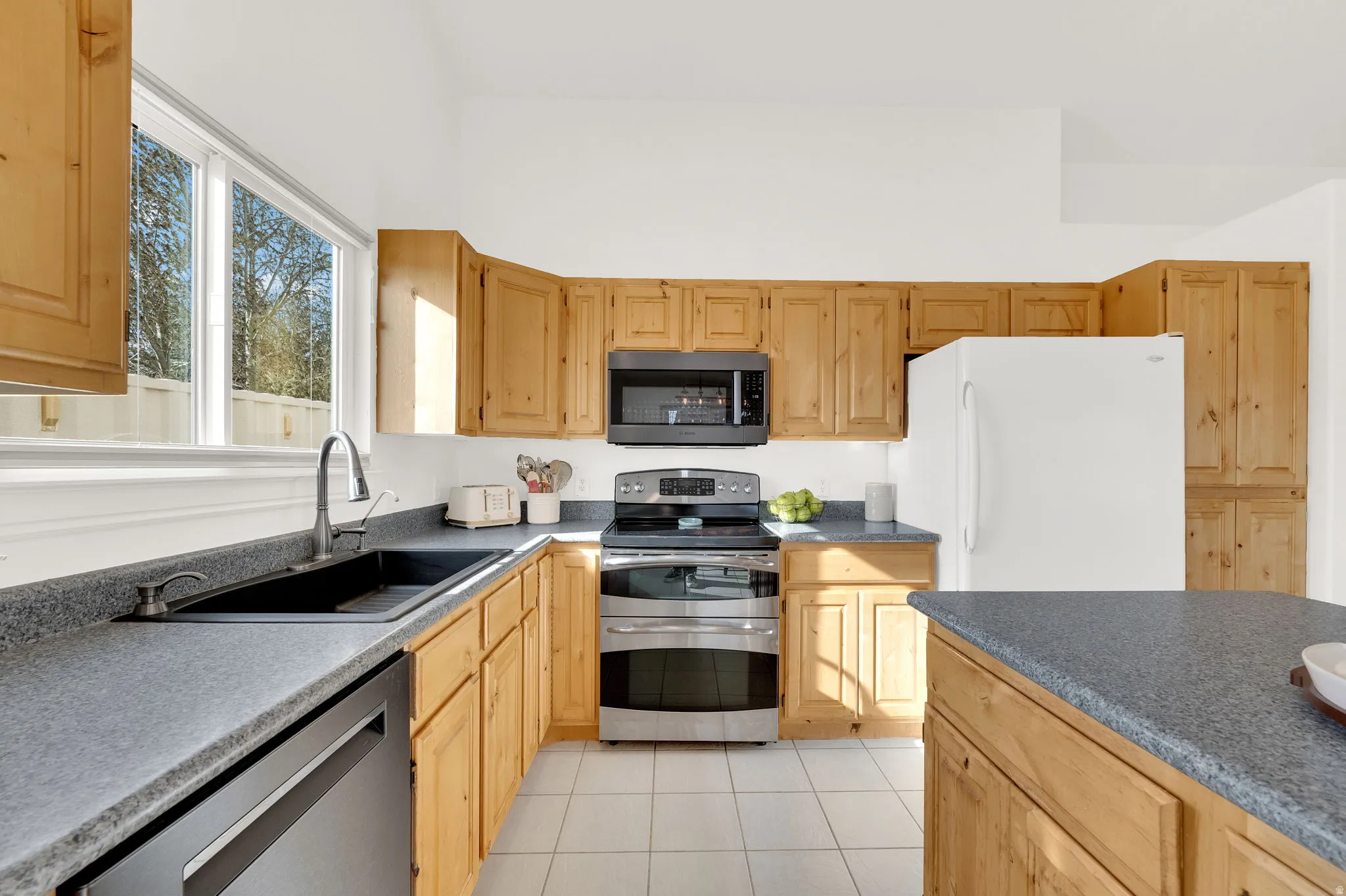 Kitchen with stainless steel appliances, dark countertops, light wood finish cabinetry, and light tile patterned floors