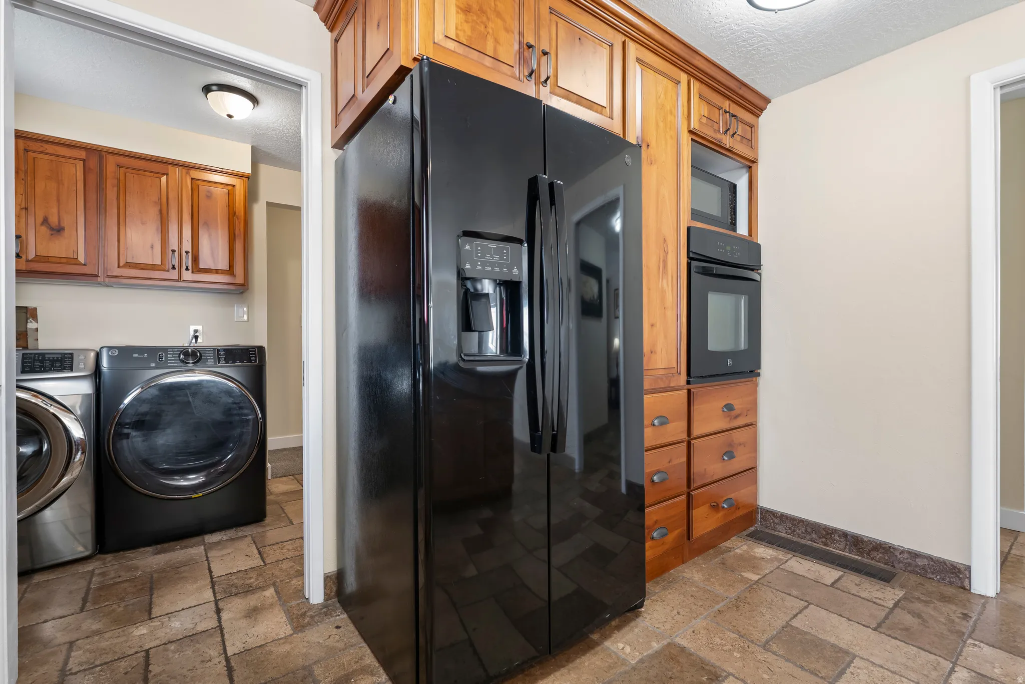 Kitchen with black appliances, a textured ceiling, wood finish cabinets, stone tile floors, and washing machine and dryer