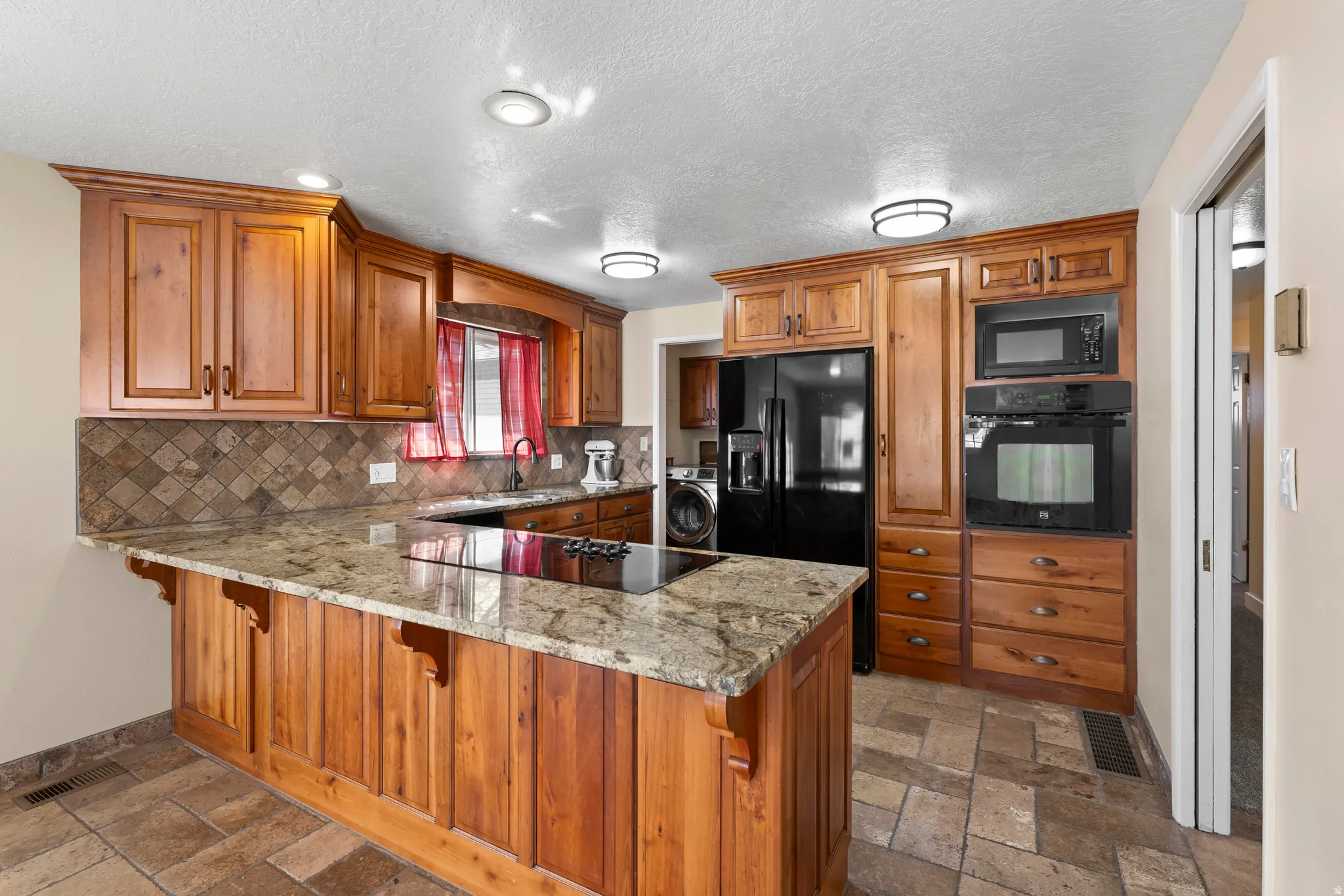 Kitchen with a peninsula, black appliances, light stone countertops, wood finish cabinetry, and a kitchen breakfast bar