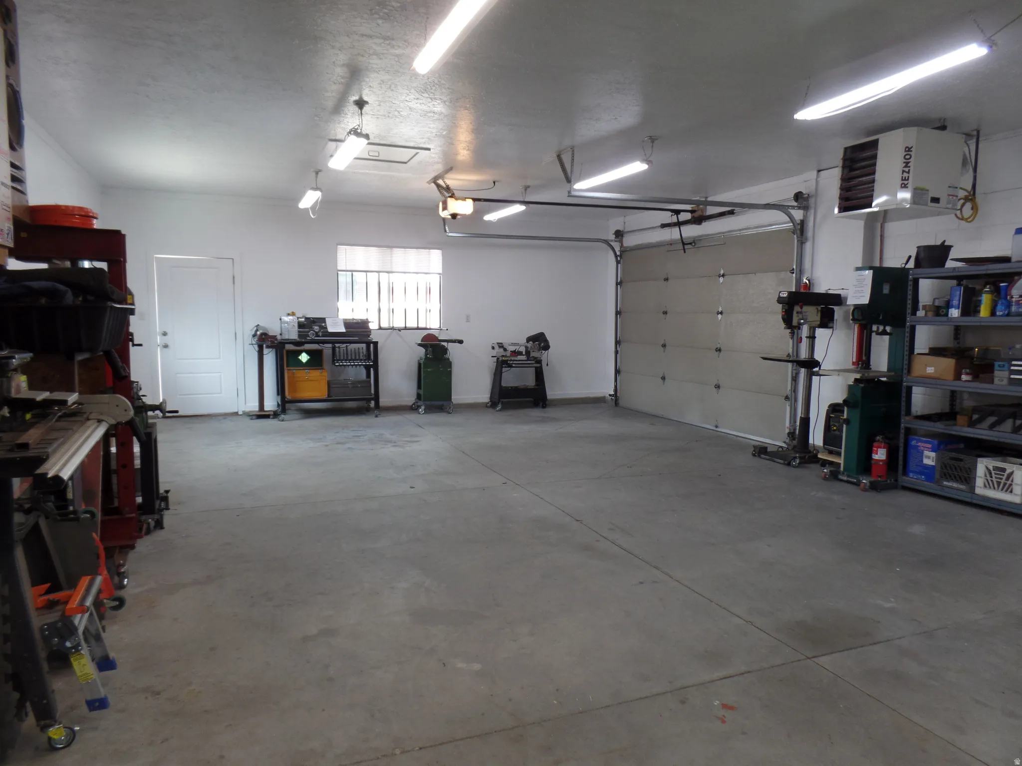 LARGE Garage featuring a workshop area, a heating unit, a 220V outlet, plumbed for air, and cabinets Spacious concrete-floored garage featuring multiple LED light fixtures, a wall-mounted heater, and extensive built-in shelving and cabinetry