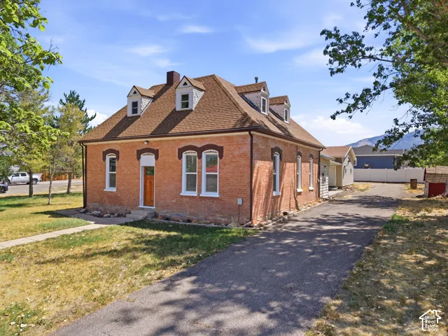 Brick exterior with dormer windows, a brown shingle roof, a wood-finish front door, and a paved driveway