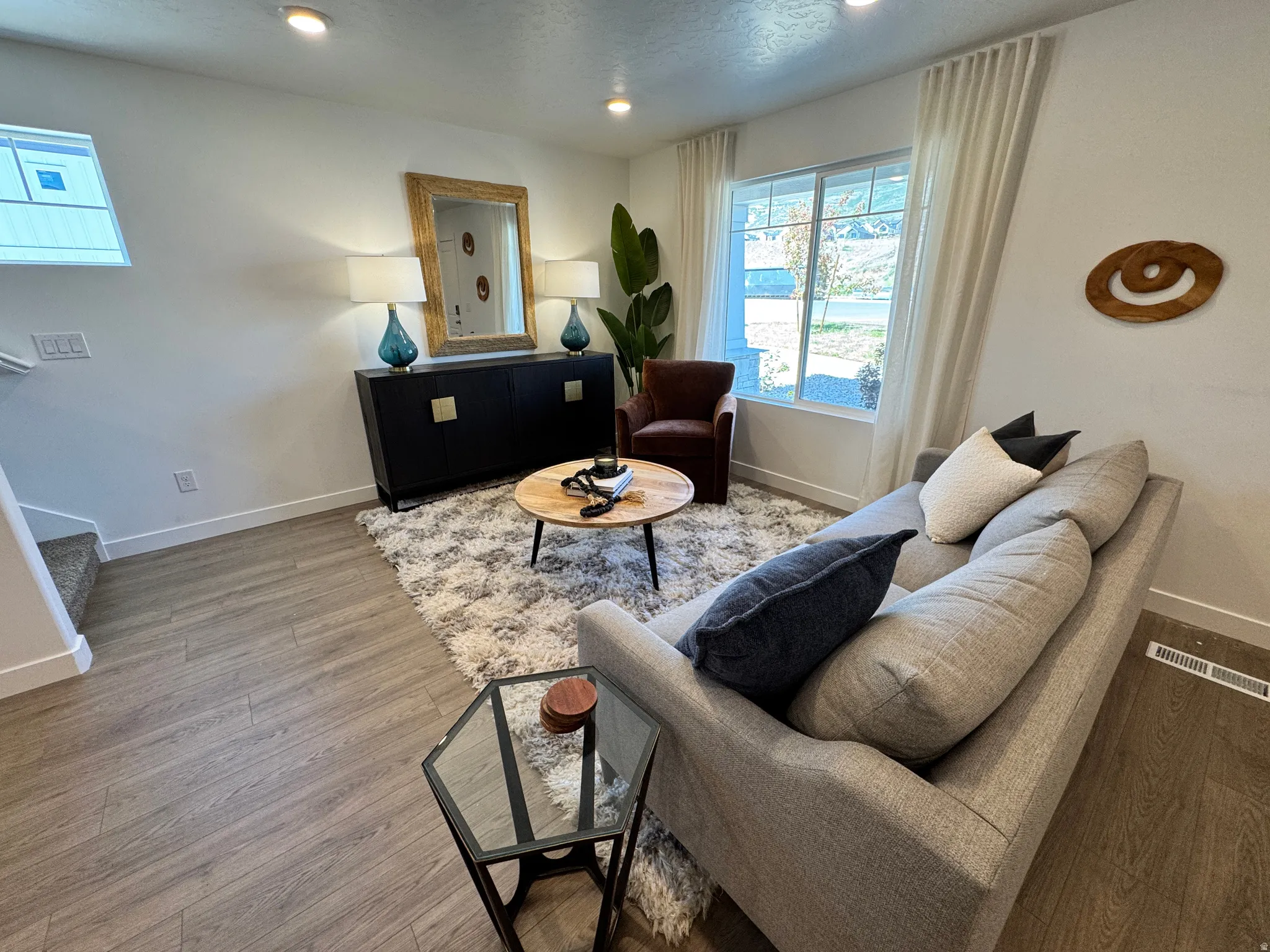 Bright living area featuring wood-finish flooring, recessed lighting, and a large window with white drapery