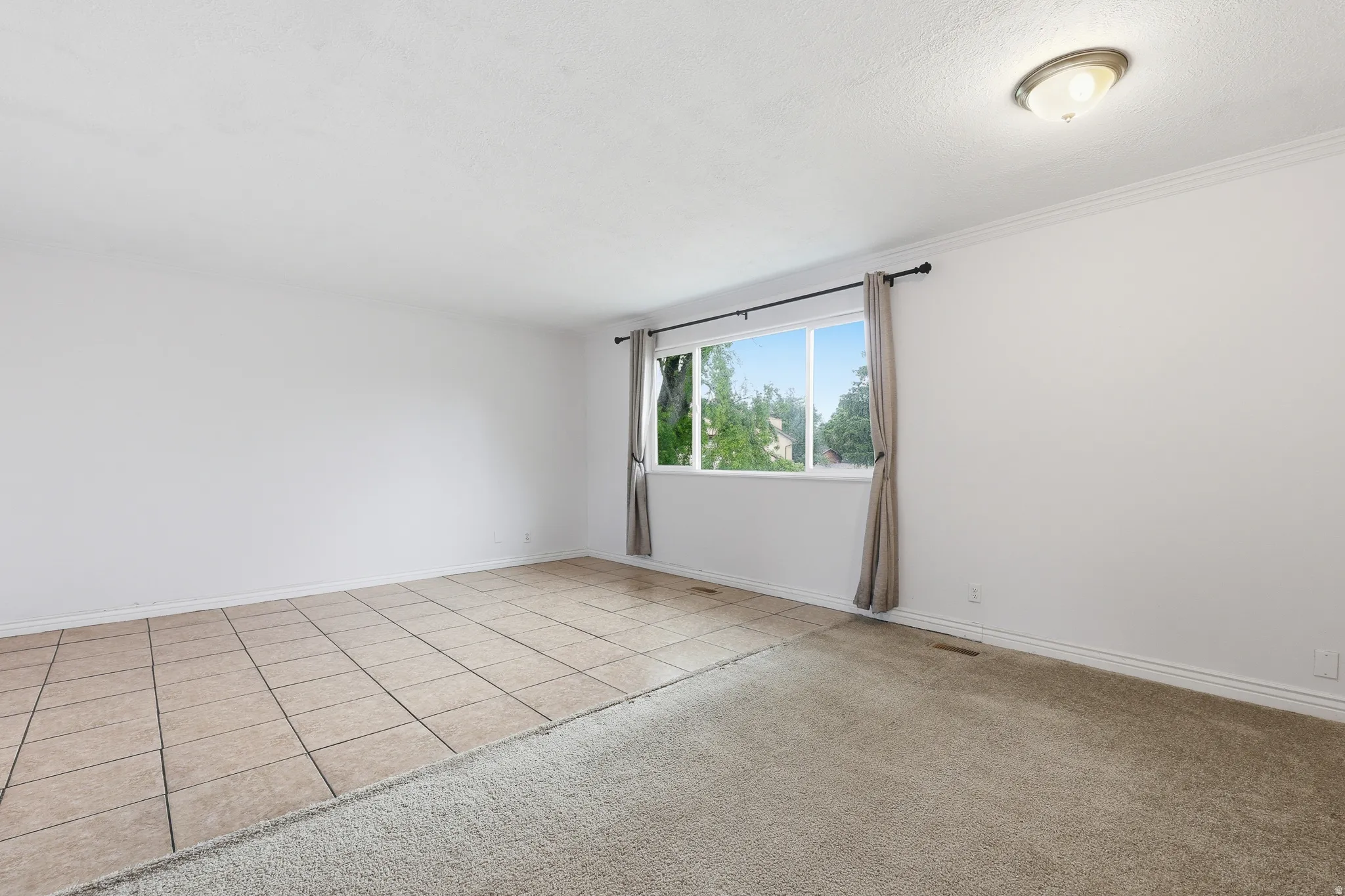 Spacious room featuring a large window, mixed flooring with ceramic tile and carpet, crown molding, and an overhead flush-mount light fixture