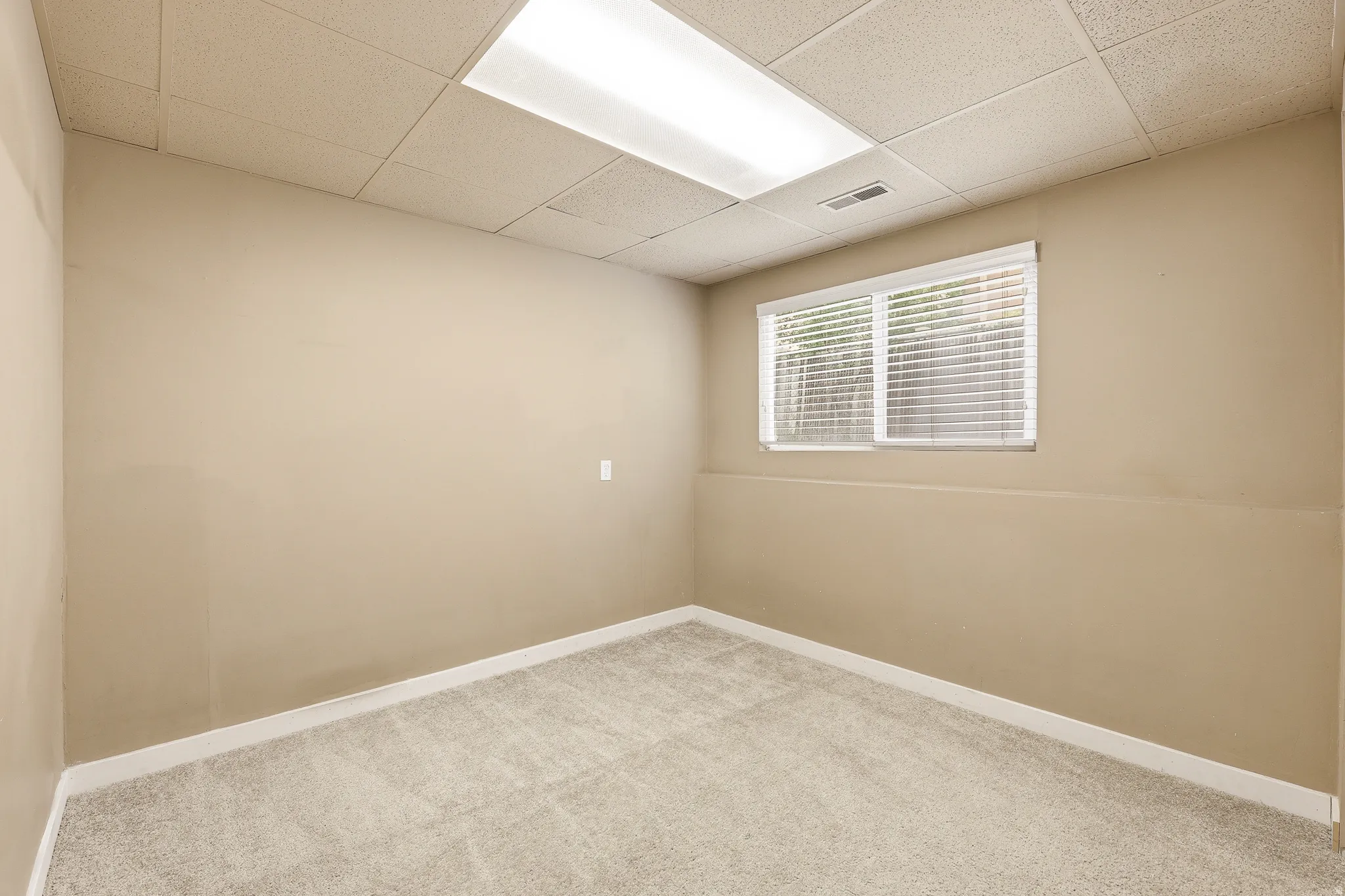 Carpeted room featuring a drop ceiling with integrated lighting, a window with blinds, and neutral painted walls with white trim