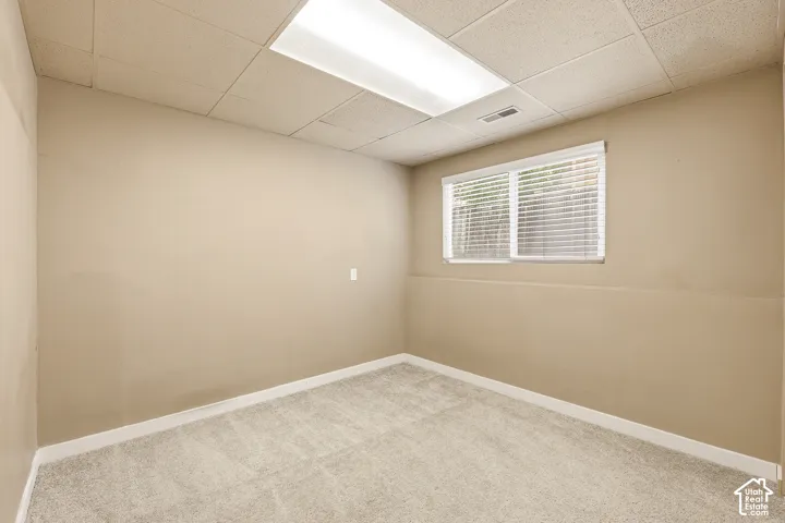 Carpeted room featuring a drop ceiling with integrated lighting, a window with blinds, and neutral painted walls with white trim