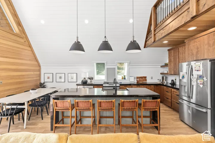 Kitchen with a center island with sink, stainless steel fridge with ice dispenser, pendant lighting, light wood-style flooring, and wooden walls