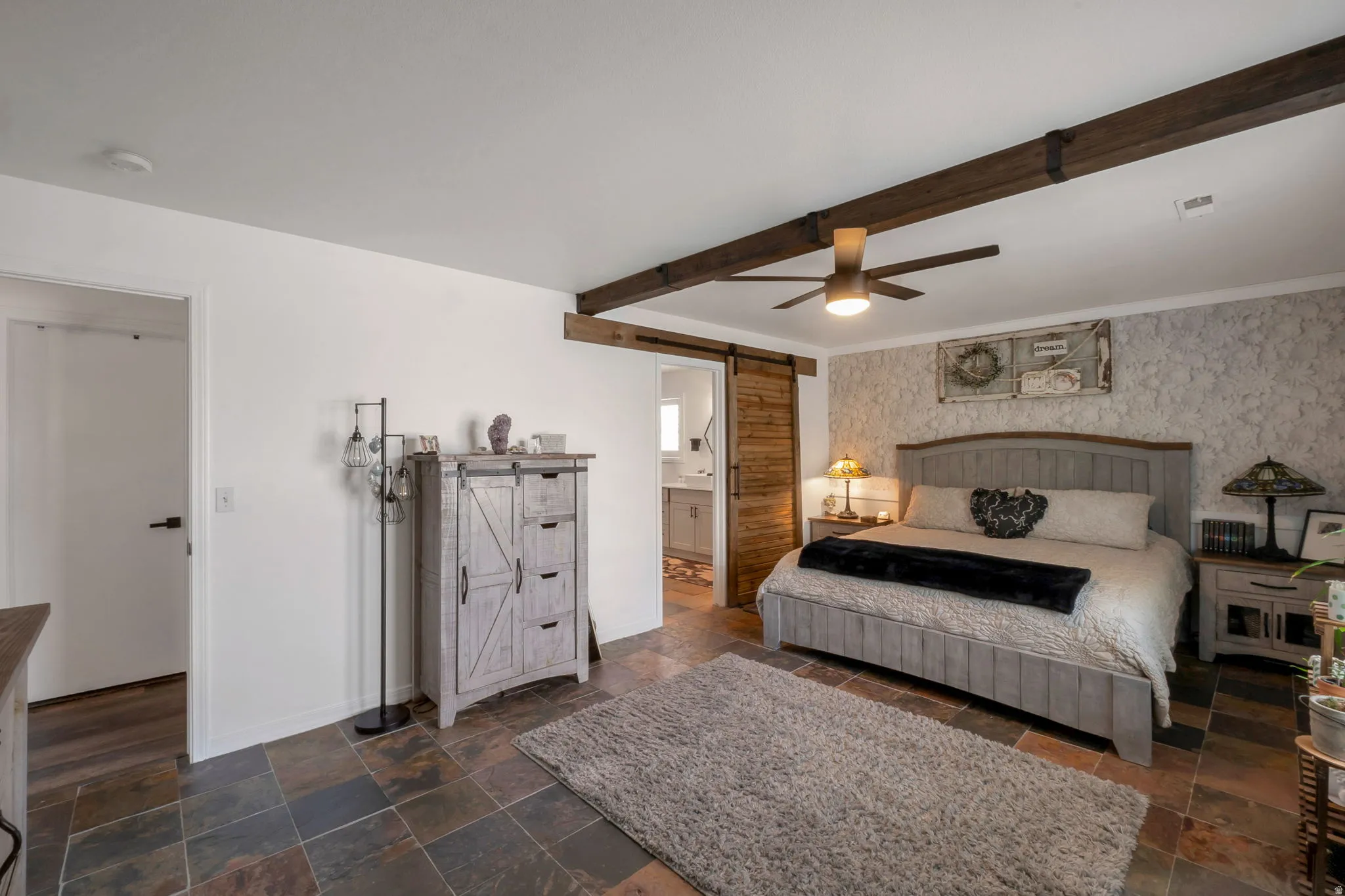 Bedroom featuring a barn door, wallpapered walls, stone tile floors, ensuite bathroom, and a ceiling fan