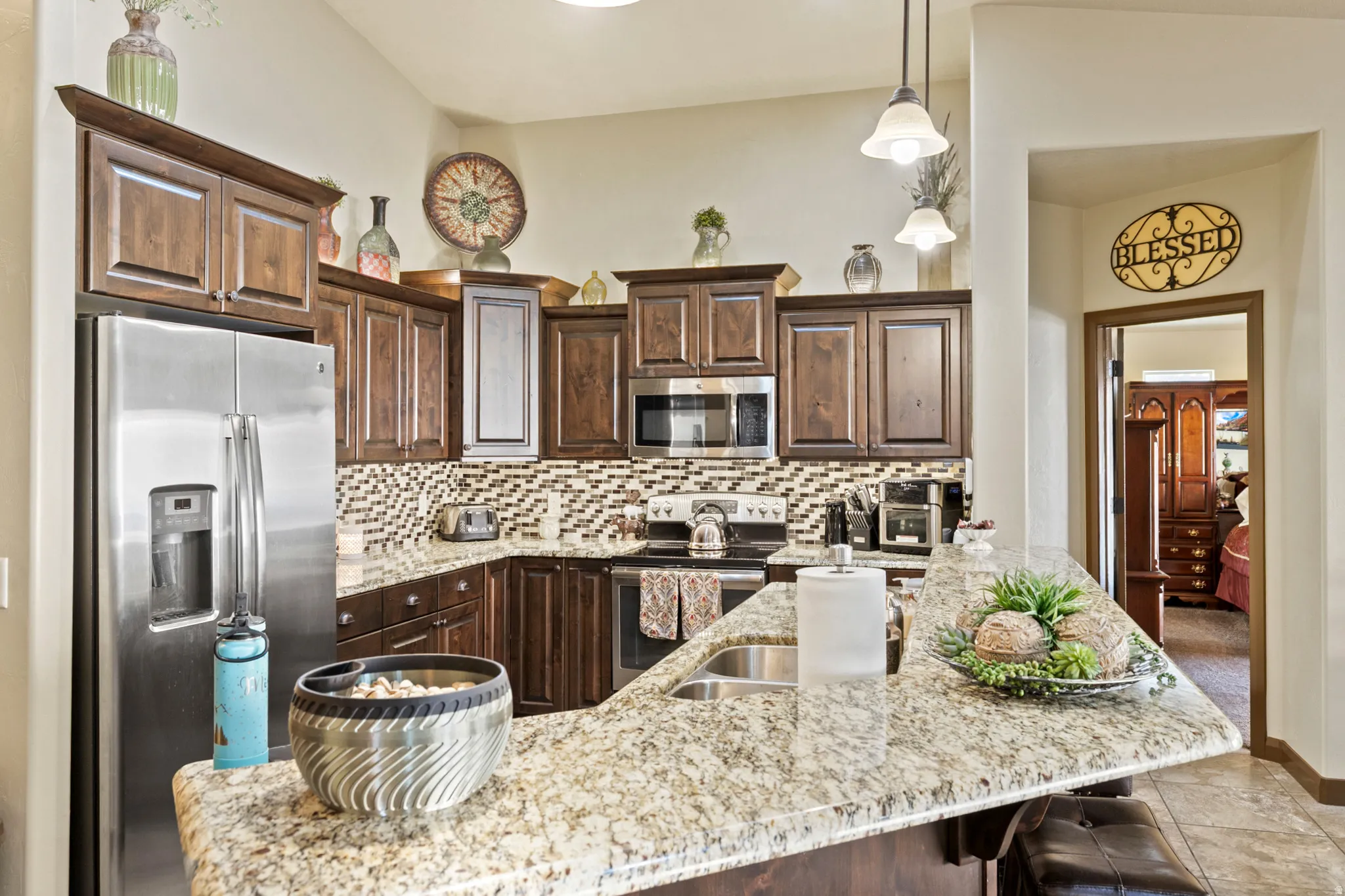 Kitchen with stainless steel appliances, dark wood finish cabinets, light stone counters, decorative light fixtures, and tasteful backsplash