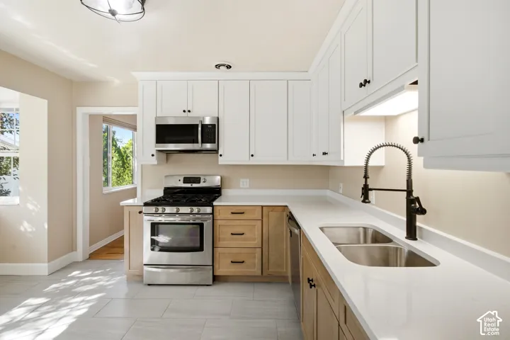 Contemporary kitchen featuring white upper cabinetry, wood-finish lower cabinetry, light-toned countertops, stainless steel appliances, and a double basin sink with a matte black faucet