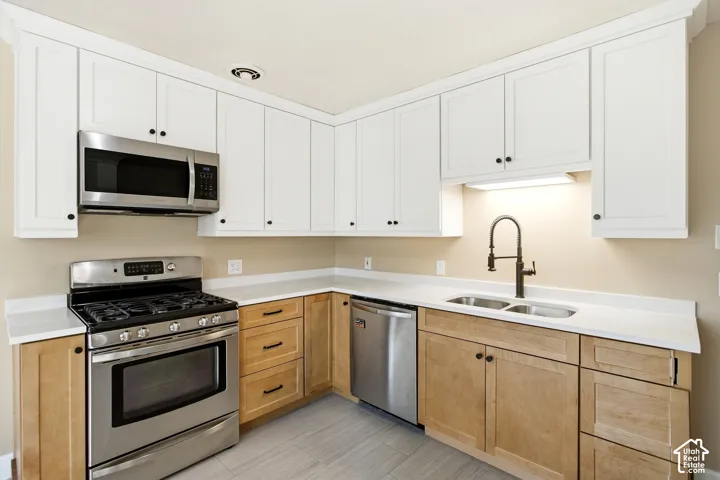 Two-tone cabinetry with white upper and wood-finish lower cabinets, stainless steel appliances, dual basin sink, wall-mounted faucet, and light-toned countertop