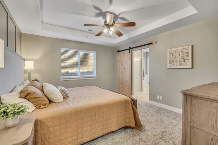 Bedroom with light colored carpet, a barn door, a raised ceiling, ceiling fan, and ensuite bathroom